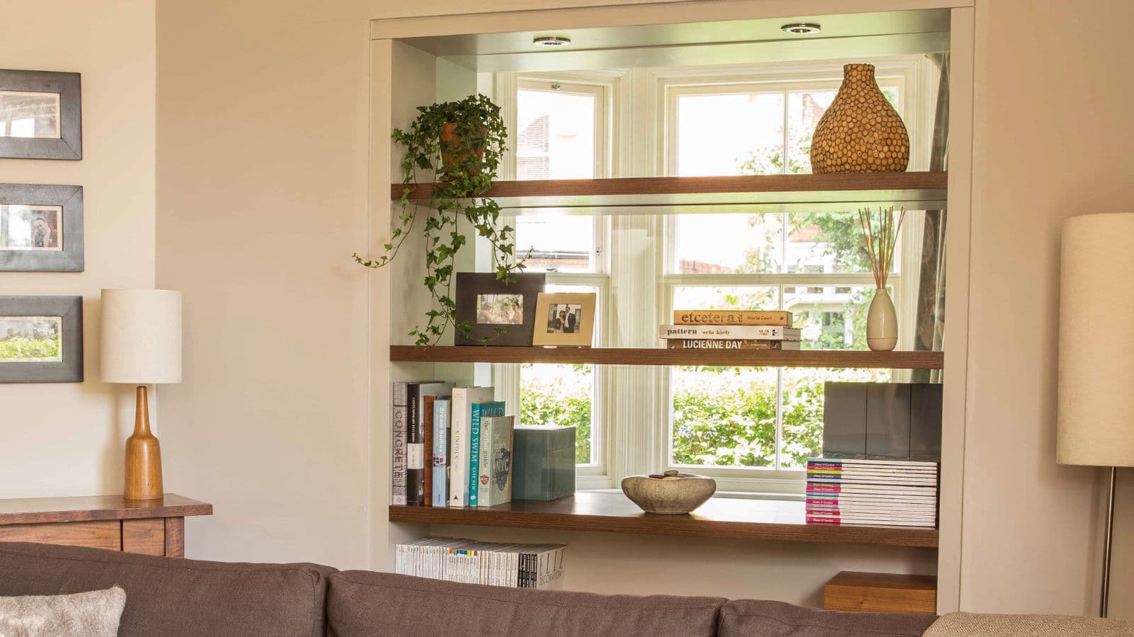 An open shelved unit built into a wide opening between two rooms, holding books and objects, with light coming through from the window beyond.
