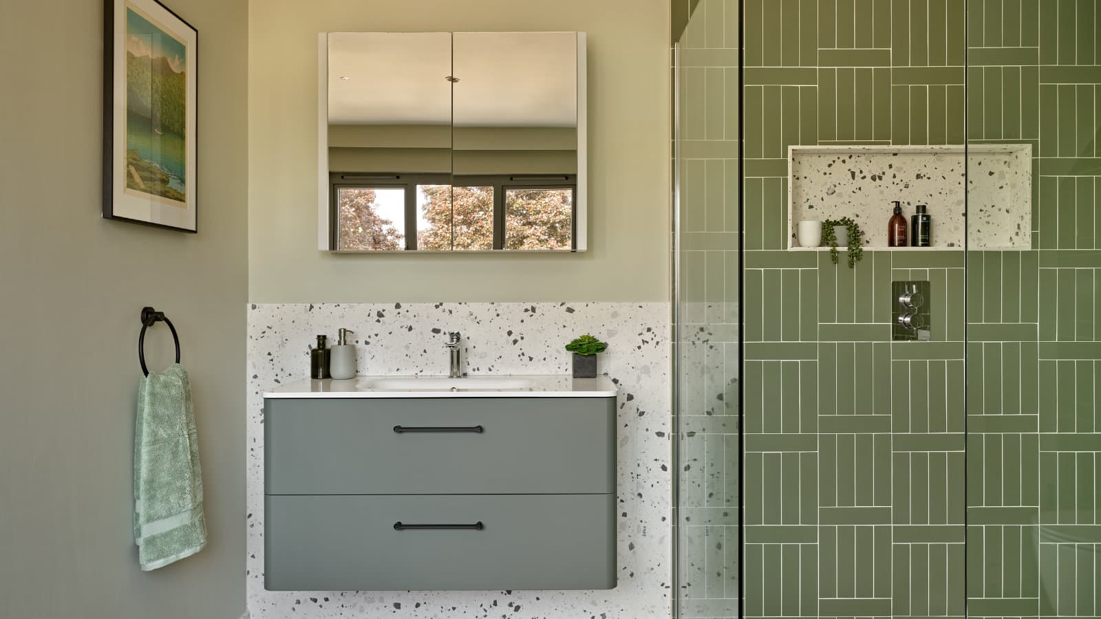 A bathroom with green vertical tiles in the shower, a terrazzo niche and splashback, a wall-hung grey vanity with black handles, and a mirrored cabinet above.